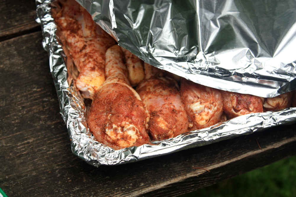 Seasoned chicken drumsticks being sealed up in a disposable aluminum pan.