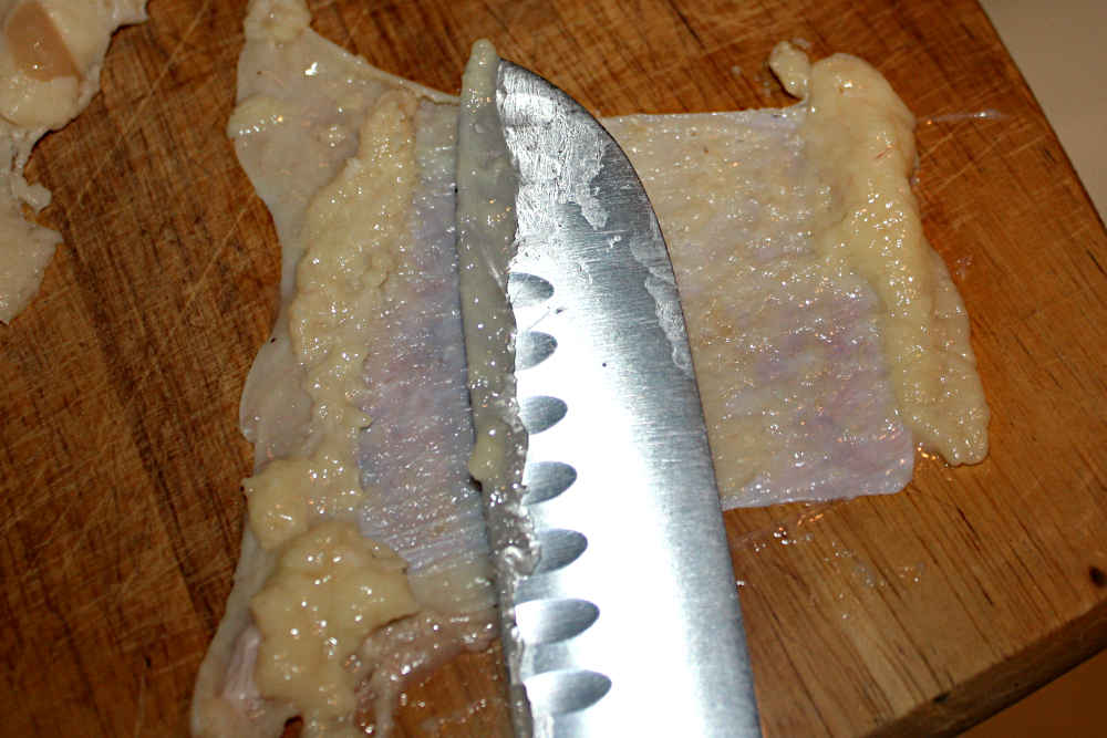 The edge of a kitchen knife scraping the fat from the underside of chicken thigh skin.